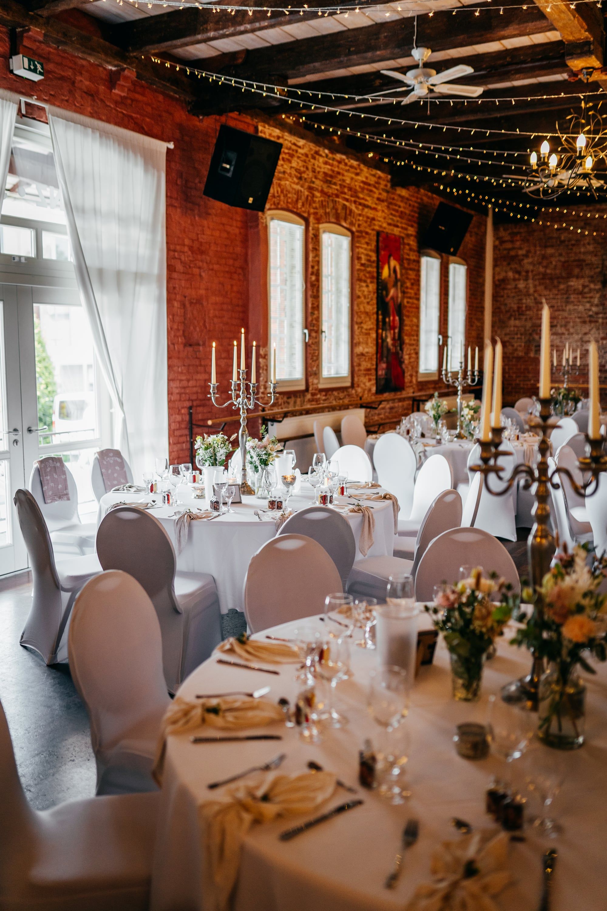 Saal mit Backsteinwand, Fenstertüren und Lichterketten, eingedeckt für eine Hochzeit