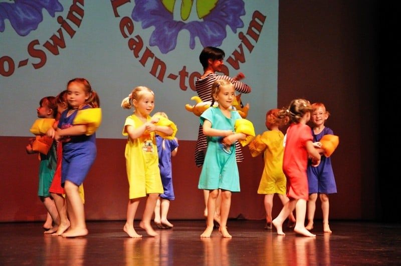 Kindergruppe mit Schwimmflügeln vor einer Kulisse mit der Aufschrift „Learn to Swim“