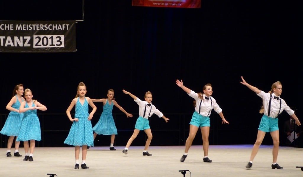 Steptanzgruppe in türkisen Kleidern und weißen Hemden mit Hosenträgern vor dem Banner der Deutschen Meisterschaft Steptanz 2013