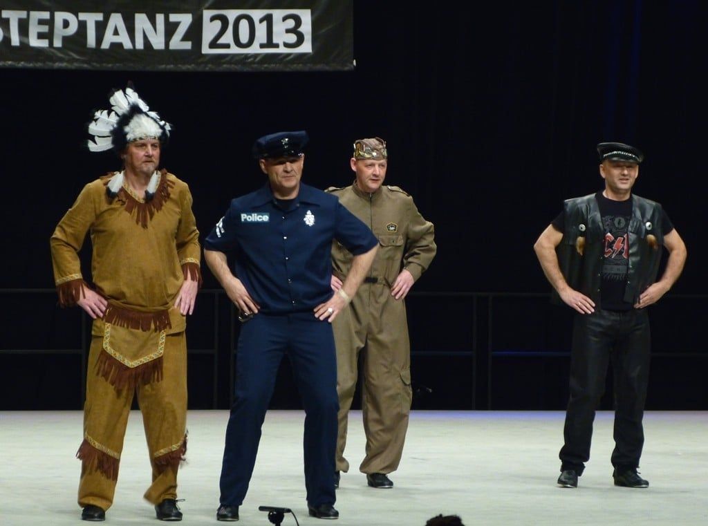Vier Tänzer in Kostümen mit Federschmuck, Polizeiuniform, Overall und Lederweste vor einem Steptanz-Banner von 2013